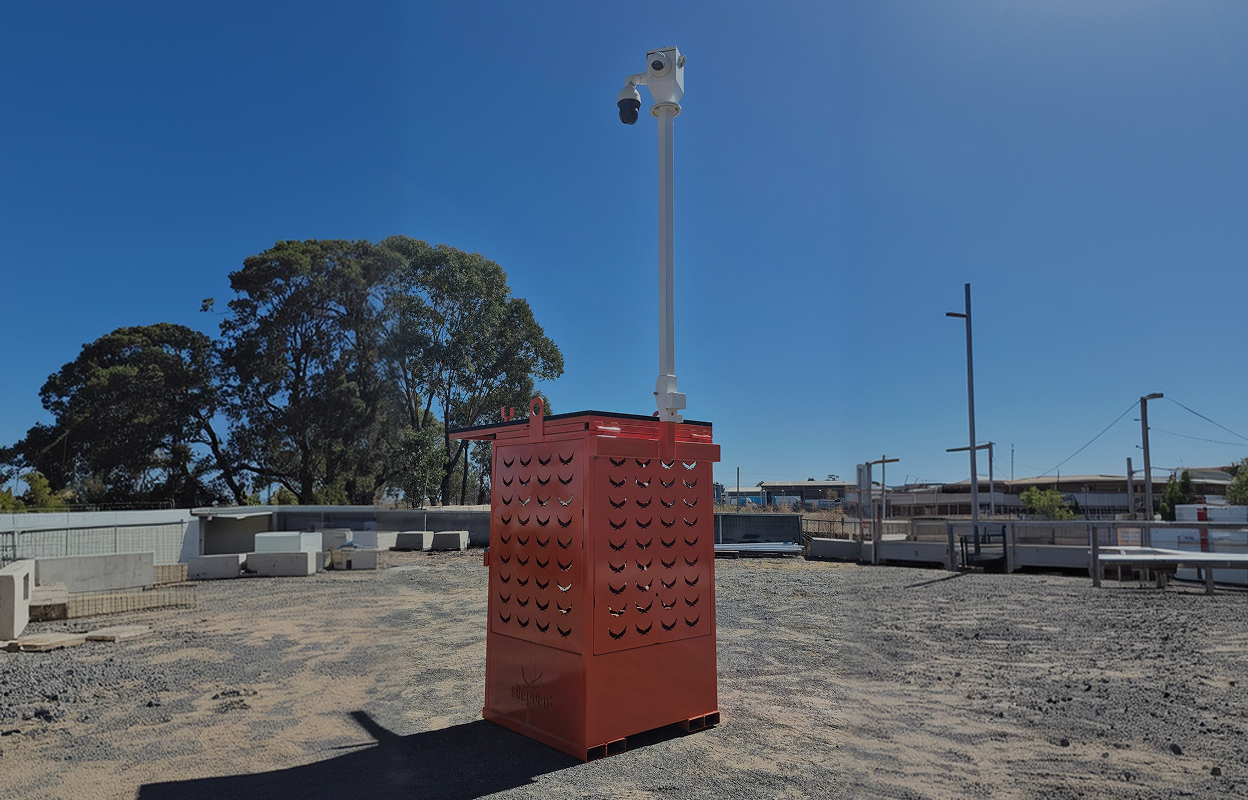 Red metal box with ventilation cutouts and a tall white pole holding a security camera, located outdoors on gravel with trees and buildings in the background under clear blue sky.